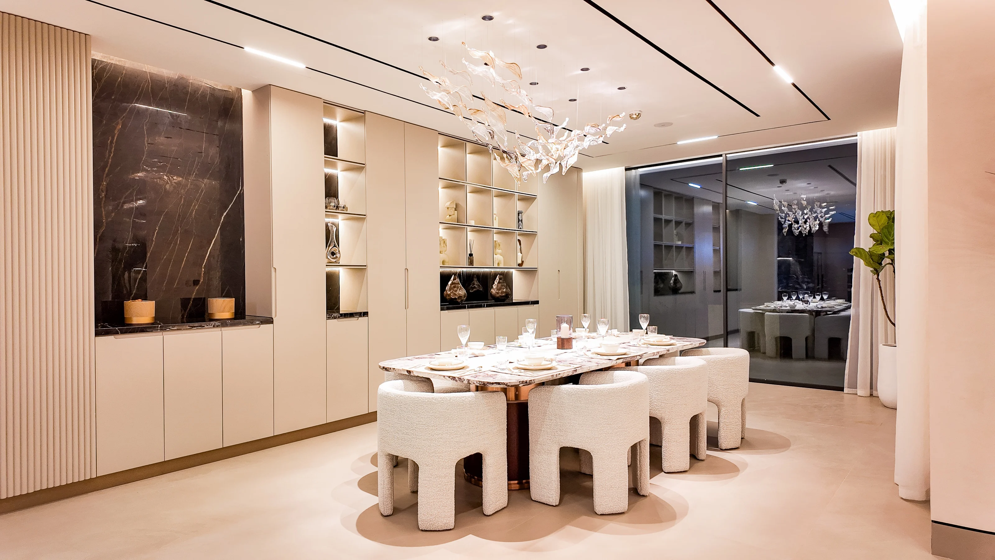 Dining room with a stone table, boucle chairs and a glass-leaf chandelier, dark marble niche beyond
