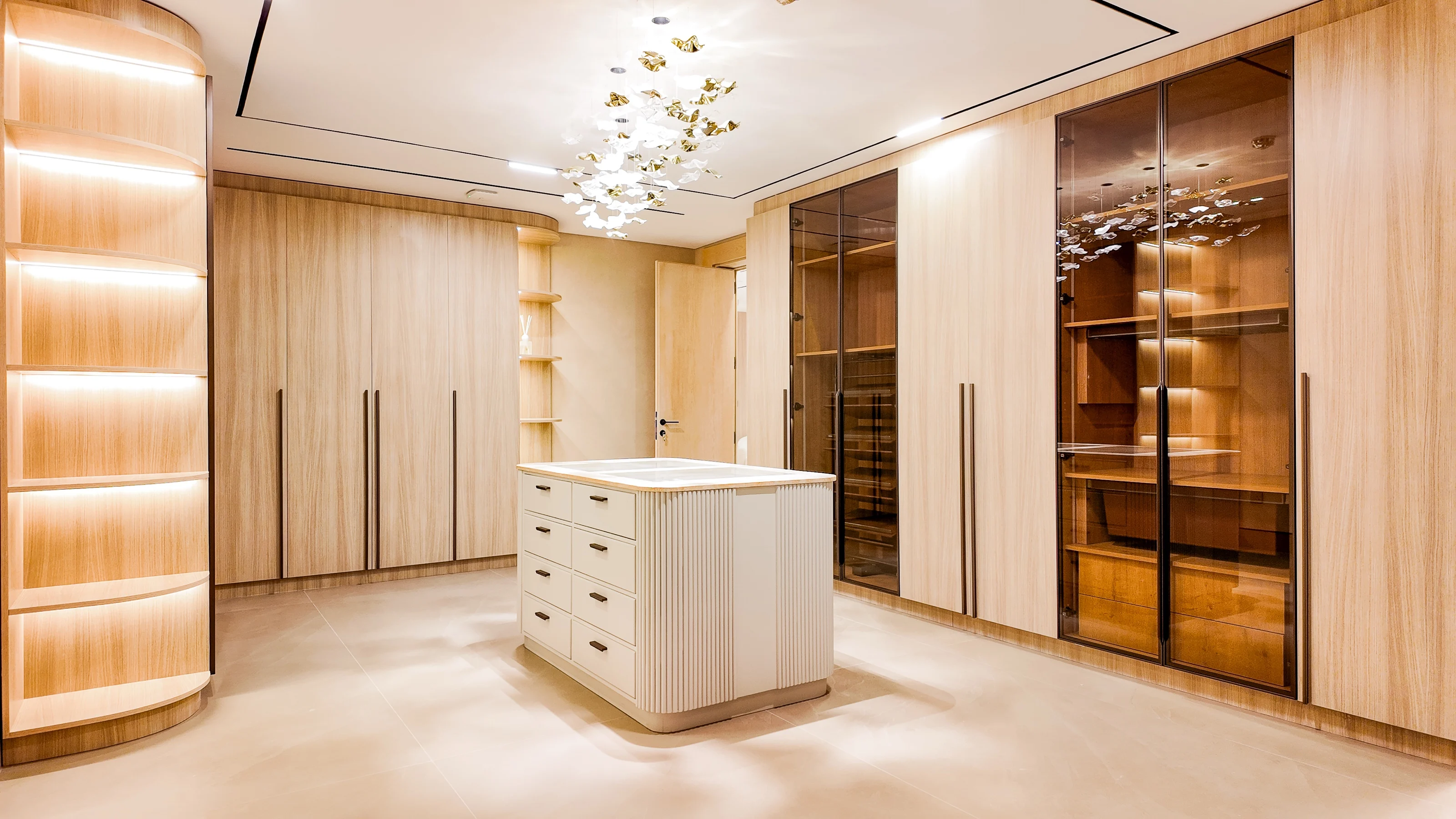 Walk-in dressing room in pale timber joinery with glass display cabinets and a gilt-leaf chandelier